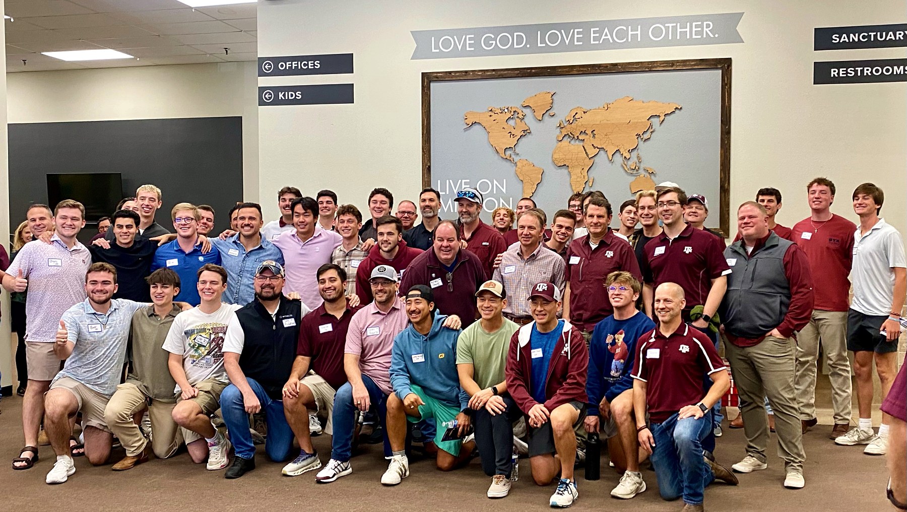A large group of BYX actives and alumni together in a church lobby beneath a sign reading Love God. Love Each Other.
