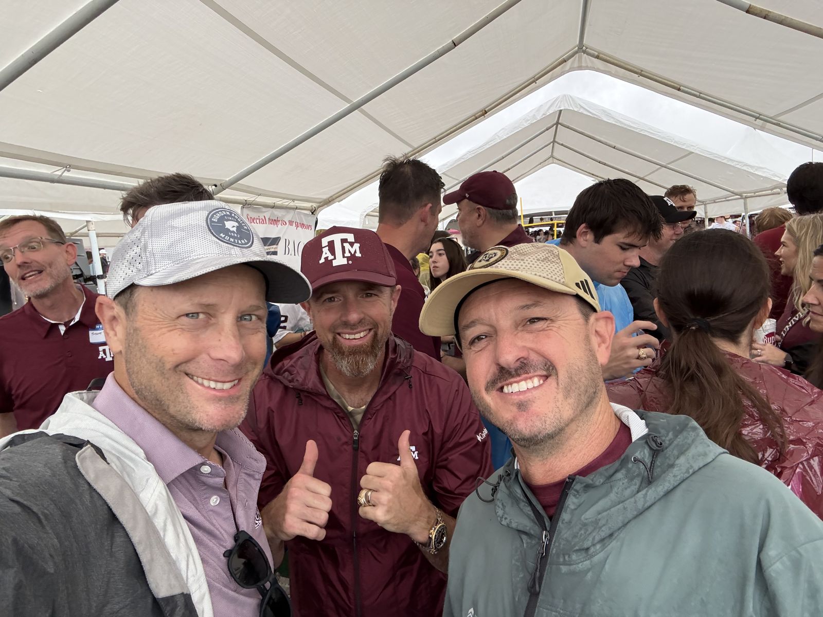 Three alumni in Aggie caps giving a thumbs up under the tent