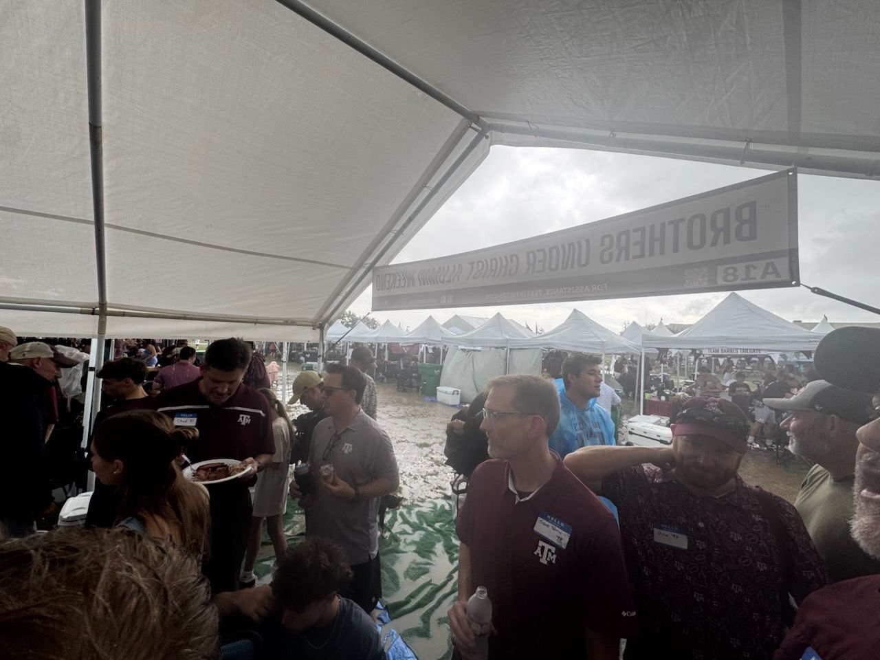 Crowd under the tent with the Brothers Under Christ banner and the muddy lawn behind